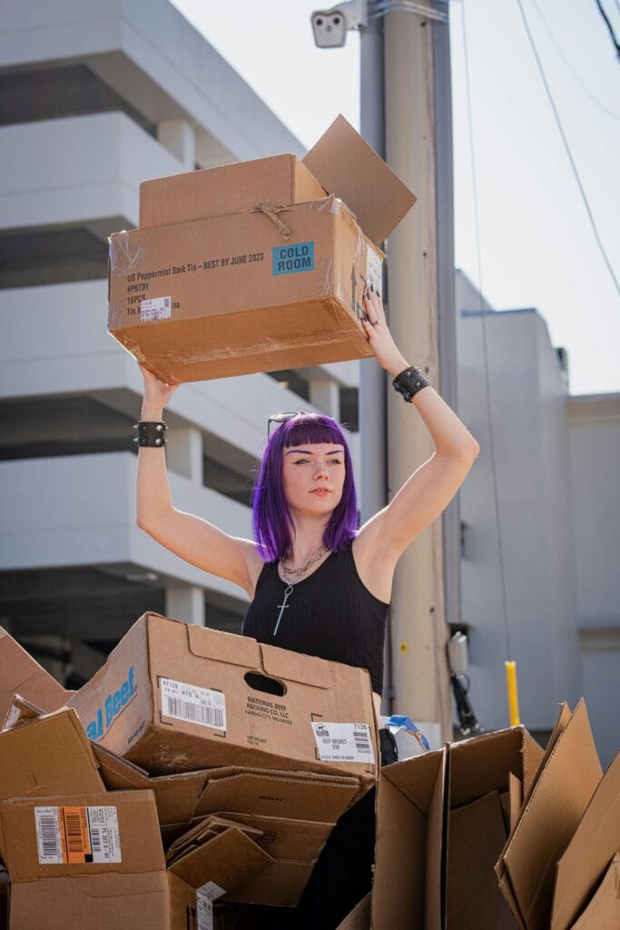 Puzzles books. A young woman with striking purple hair and gothic accessories lifts a cardboard box above her head while standing among a pile of flattened boxes in an urban alley, with modern buildings in the background and sunlight casting strong shadows.