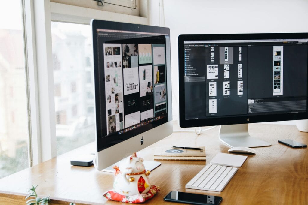Featuring Spot the Difference Puzzle design. Workspace with two large desktop monitors displaying design layouts, a white keyboard, mouse, smartphone, notebook, and a ceramic lucky cat figurine on a wooden desk near a window.