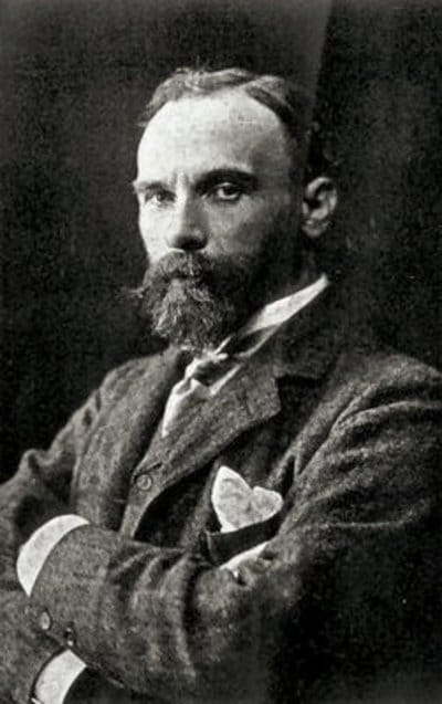 Black and white portrait of a man with a serious expression. He has a beard, wears a suit and tie, and stands with arms crossed, conveying a confident tone.